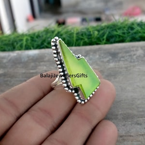 May include: A silver ring with a large, light green, geometric gemstone. The gemstone is shaped like a lightning bolt. The ring has a beaded border and the text "BalajiJewellersGifts".