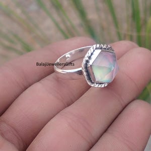 May include: A silver ring with a large, faceted, iridescent gemstone. The gemstone has a pale pink and green hue. The ring band is smooth, while the setting around the stone has a textured, rope-like design. The text "BalajiJewellersGifts" is visible.