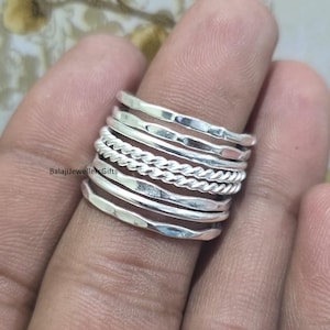 Può includere: Un set di anelli impilabili in argento con più fasce con design strutturati e intrecciati. Gli anelli sono mostrati su un dito, mostrando il loro aspetto a strati. Il testo "BalajiJewellersGifts" è visibile.