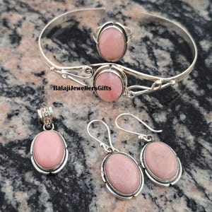 May include: A jewelry set featuring a ring, bracelet, pendant, and earrings. Each piece has a light pink oval stone set in a silver-colored metal. The bracelet and ring have a similar design, while the pendant has a decorative bail.
