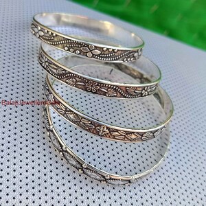 May include: Four silver bangles with intricate floral designs. The bangles are stacked on top of each other.
