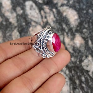 Może przedstawiać: Srebrny pierścionek z owalnym, fasetowanym rubinem. Pierścionek ma bogaty, szczegółowy wzór ze zdobieniami i koralikami. Tekst "BalajiJewellersGifts" jest widoczny z boku pierścionka.