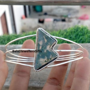 May include: A silver bangle bracelet featuring a triangular moss agate stone. The stone is set within a silver frame with a twisted rope design. The bracelet has three parallel bands. The text "BalajiJewellersGifts" is visible.