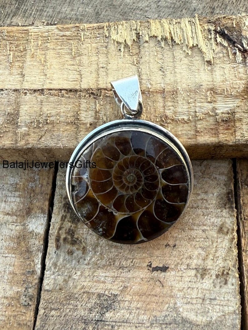May include: A silver pendant featuring a round ammonite fossil with a brown and white spiral pattern. The pendant is on a wooden surface.