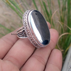 Pode incluir: Um anel de prata com uma pedra oval cinza escuro. O anel tem um design ornamentado e detalhado ao redor da pedra e na banda. O texto "BalajiJewellersGifts" é visível no anel.