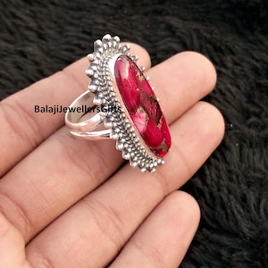 May include: A silver ring featuring a large, oval, red gemstone. The gemstone has a textured appearance with gold flecks. The ring's setting has a detailed, beaded design around the stone and on the band. The text "BalajiJewellersGifts" is visible.