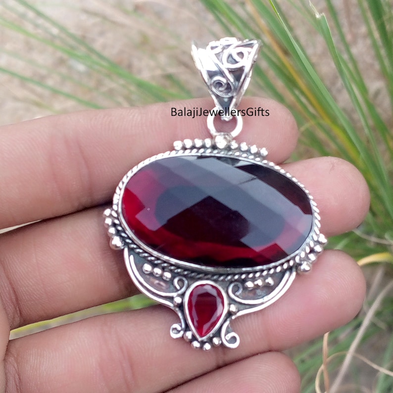 May include: A silver pendant with a large, faceted, oval-shaped, deep red gemstone as the focal point. A smaller, teardrop-shaped red stone is below. The pendant has ornate silver detailing and a decorative bail. The text "BalajiJewellersGifts" is visible.