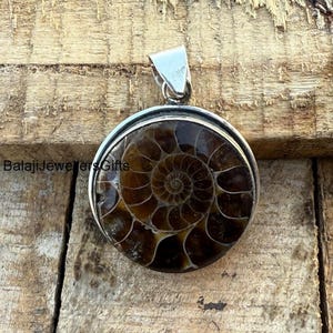 May include: A silver pendant featuring a round ammonite fossil with a brown and white spiral pattern. The pendant is on a wooden surface.