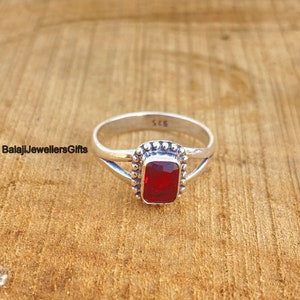 May include: A silver ring with a red gemstone set in a square bezel. The ring has a delicate, criss-cross design on the band. The ring is marked "925" on the inside.