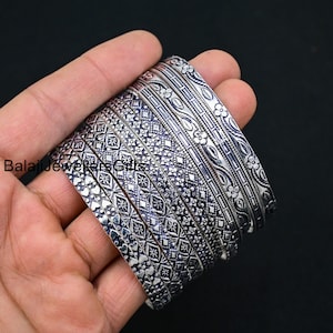 May include: A stack of silver bangles with intricate floral and geometric patterns. The bangles are held in a hand against a dark background, showcasing the detailed designs and metallic sheen. The text "Balaji Jewelers Gifts" is visible.