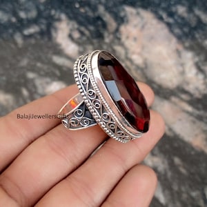 May include: A silver ring with an oval, faceted, deep red gemstone. The ring has an ornate, filigree design around the gemstone and on the band. The ring is held in a hand, with the text "BalajiJewellersGifts" visible on the band.