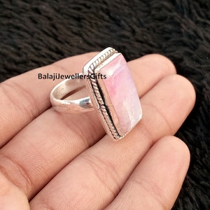 May include: A silver ring featuring a rectangular, pale pink gemstone. The ring has a textured border and the text "BalajiJewellersGifts" is visible. The ring is held in a hand.