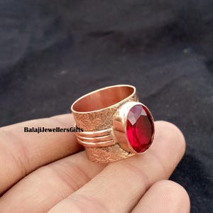 Puede incluir: Un anillo de color cobre con una piedra preciosa ovalada de color rojo intenso. El anillo tiene un patrón floral grabado en la banda y el texto "BalajiJewellersGifts". La piedra preciosa es el punto focal, engastada en un bisel de cobre pulido.