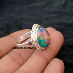 Puede incluir: Un anillo de plata con una piedra de ópalo en forma de pera. El ópalo muestra un vibrante juego de colores, con tonos rojos, verdes y azules. El anillo tiene un engaste detallado y una banda lisa. El texto "BalajiJewellersGifts" es visible.
