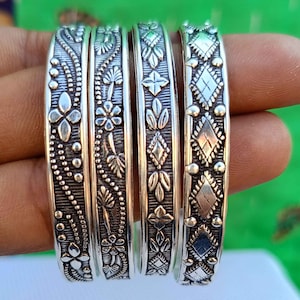 May include: Four silver bangles with intricate floral and geometric designs. The bangles are stacked on top of each other and have a shiny finish.