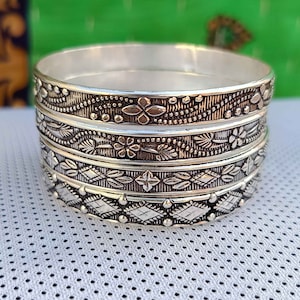 May include: Four silver bangles with intricate floral and geometric designs. The bangles are stacked on top of each other and have a shiny finish.