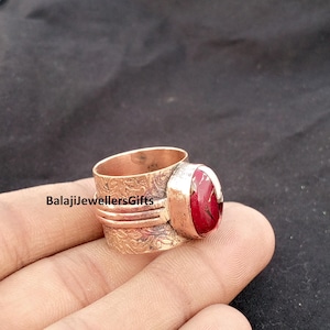 May include: A wide copper ring featuring an embossed floral pattern and a central oval-shaped red stone with dark veining. The band has a textured finish and the text BalajiJewellersGifts is visible on the surface of the metal ring band.