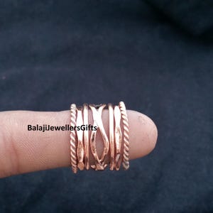 May include: A copper-coloured spinner ring with multiple bands, including twisted rope and smooth designs. The ring is displayed on a finger against a dark background. The text "BalajiJewellersGifts" is visible.