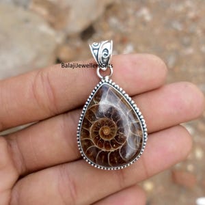 May include: A teardrop-shaped pendant with a fossilized ammonite encased in a silver-colored setting. The fossil displays a spiral pattern in shades of brown and tan. The pendant has a decorative bail and the text "BalajiJewellersGifts".