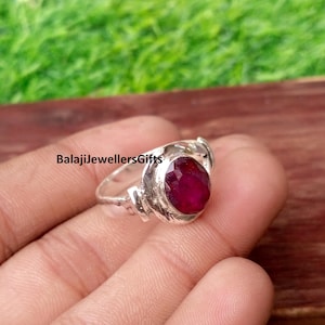 May include: A silver ring featuring a faceted, deep red gemstone set in a textured metal band. The ring displays the text BalajiJewellersGifts on the surface. The metal band has a polished finish with visible detailing around the central stone setting.