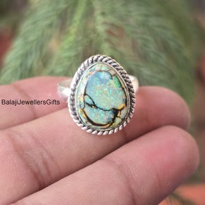 May include: A silver ring featuring an oval-shaped, iridescent gemstone with a unique cracked pattern. The gemstone displays shades of green, blue, and gold. The ring's band has a twisted rope design around the gemstone. The text "BalajiJewellersGifts" is visible.