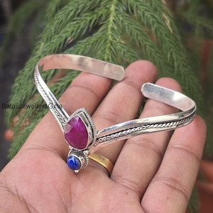 Könnte beinhalten: Ein silbernes Armreif-Armband mit einem tropfenförmigen, magentafarbenen Edelstein und einem kleineren, runden, blauen Edelstein. Das Armband hat ein strukturiertes Design an den Seiten. Der Text "Balaji Jewellers Gifts" ist sichtbar.