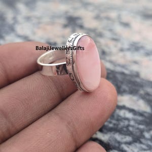 May include: A silver ring with a large, oval, light pink gemstone. The band is plain, while the setting around the stone has a decorative, etched design. The ring is held in a hand. The text "BalajiJewellersGifts" is visible.