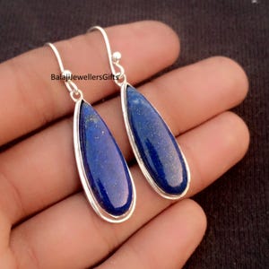 May include: A pair of teardrop-shaped earrings with a deep blue stone set in a silver metal frame. The earrings have a hook closure. The text "BalajiJewellersGifts" is visible on the left earring.