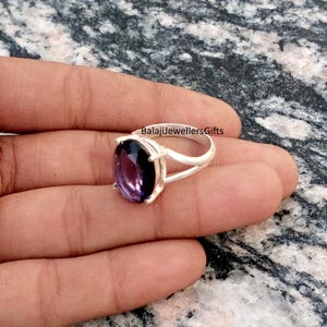 May include: A silver ring with a large, oval-cut amethyst gemstone. The amethyst is a deep purple color. The ring has a split shank design and the text "BalajiJewellersGifts" is visible.