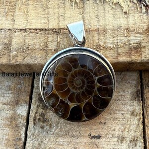 May include: A silver pendant featuring a round ammonite fossil with a brown and white spiral pattern. The pendant is on a wooden surface.