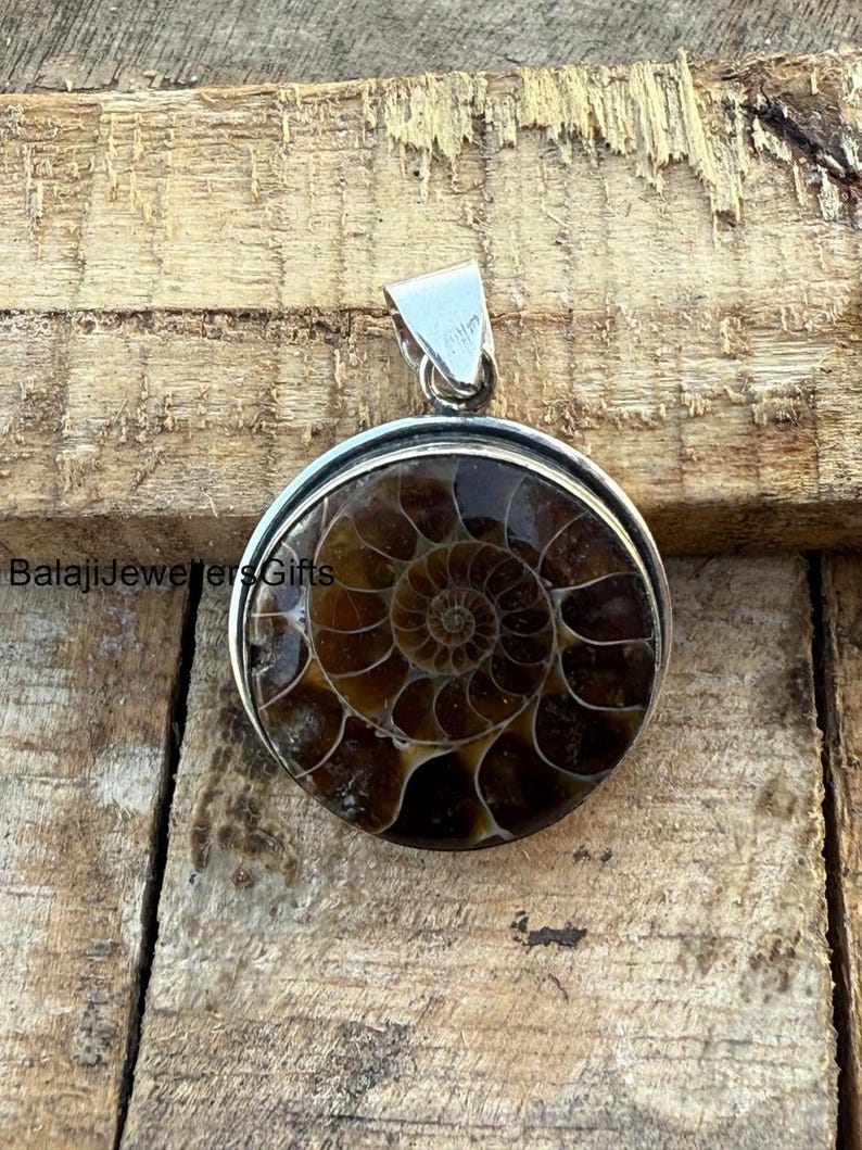 May include: A silver pendant featuring a round ammonite fossil with a brown and white spiral pattern. The pendant is on a wooden surface.