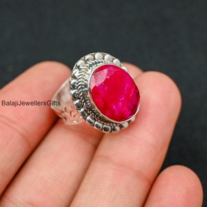 May include: A silver ring with a large, oval-shaped, bright red ruby gemstone set in a silver bezel. The ring has a decorative, textured band.