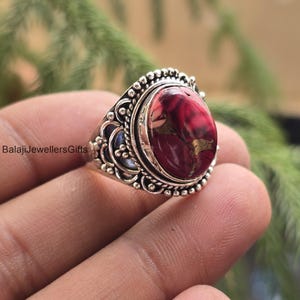 Puede incluir: Anillo de plata con una piedra ovalada roja y dorada. La piedra tiene una textura única. El anillo presenta detalles intrincados alrededor de la piedra y la banda. Una joya llamativa.