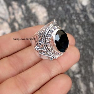 Può includere: Un anello d'argento con una pietra nera ovale. L'anello presenta un design ornato con dettagli floreali e a volute. Il testo "BalajiJewellersGifts" è visibile sul lato dell'anello.