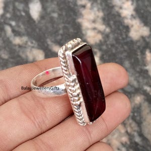 Puede incluir: Un anillo de plata con una piedra preciosa rectangular de color rojo oscuro. El anillo tiene un borde texturizado y una banda lisa. El texto "BalajiJewellersGifts" es visible en la banda del anillo. El anillo se sostiene en una mano.