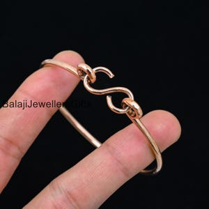 May include: A gold-toned bangle bracelet with a unique S-shaped clasp. The bracelet is made of metal and is a simple, elegant design.