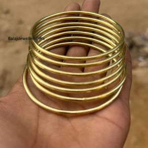 May include: A stack of gold-colored bangles, each a simple, unbroken circle. The bangles are held in a hand, with the text "BalajiJewellersGifts" visible in the background. The bangles appear to be made of metal.