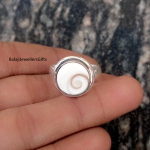 May include: A silver ring featuring a white, circular stone with a spiral design in the center. The ring has a simple band and a bezel setting. The text "BalajiJewellersGifts" is visible on the left side.