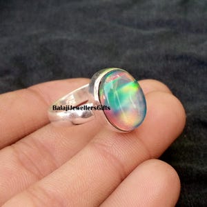 Op de afbeelding: Een zilveren ring met een ovale, iriserende edelsteen. De edelsteen toont een spectrum aan kleuren, waaronder groen, blauw en roze. De ring wordt in een hand gehouden en de tekst "BalajiJewellersGifts" is zichtbaar op de ringband.