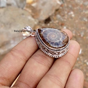 May include: A teardrop-shaped pendant with a polished, brown fossil set in a silver filigree frame. The fossil displays a spiral pattern. The pendant has a small loop at the top for a chain. The text "BalajiJewellersGifts" is visible.