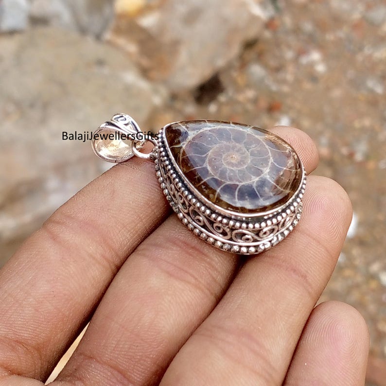 May include: A teardrop-shaped pendant with a polished, brown fossil set in a silver filigree frame. The fossil displays a spiral pattern. The pendant has a small loop at the top for a chain. The text "BalajiJewellersGifts" is visible.