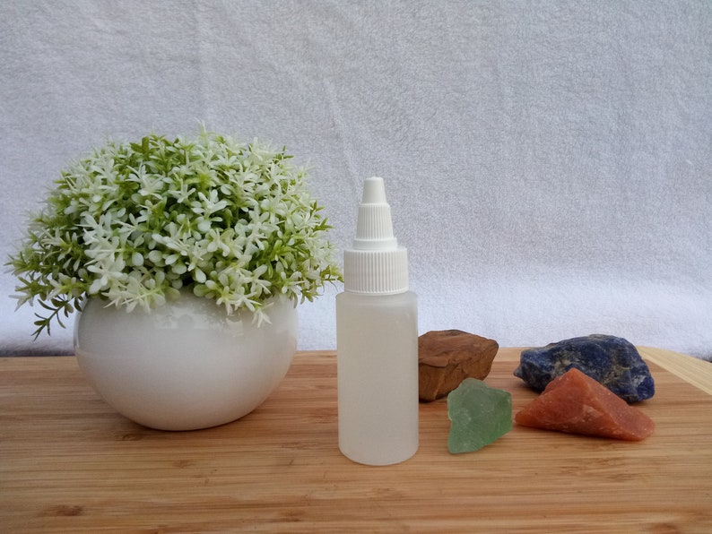 Op de afbeelding: Een doorzichtige plastic fles met een witte dop staat op een houten oppervlak, naast een witte keramische pot met een groen-witte plant. Er zijn ook verschillende gekleurde stenen aanwezig, waaronder rood, groen en blauw.