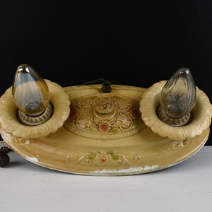 May include: Antique light fixture with two bulbs. The fixture has a cream-colored, oval-shaped base with floral and scrollwork details. The bulbs are teardrop-shaped and have a vintage appearance. A pull chain hangs from the fixture.