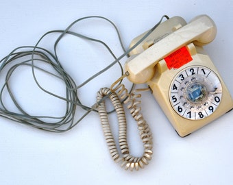 Könnte beinhalten: Cremefarbenes Vintage-Wählscheibentelefon mit Spiralschnur und geradem Kabel. Das Zifferblatt hat schwarze Zahlen und Buchstaben. Auf dem Telefon befindet sich ein roter Aufkleber. Das Telefon liegt auf einer weißen Oberfläche.