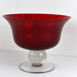 May include: A ruby red glass bowl with a clear glass base. The bowl has a wide, flared rim and a rounded body. The base consists of a spherical center and a circular foot. The bowl is likely for decorative purposes.