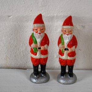 May include: Two vintage Santa Claus figurines with white beards and red and white suits. They are holding green sprigs in their hands and are standing on silver bases.