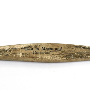 May include: A vintage, elongated wooden sign with the text "Best in Meats and Groceries" in black lettering. The sign has a weathered, aged appearance with a light brown and dark brown color scheme.
