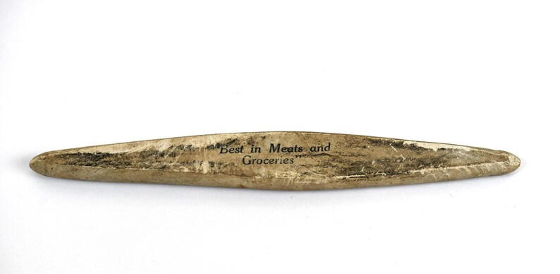 May include: A vintage, elongated wooden sign with the text "Best in Meats and Groceries" in black lettering. The sign has a weathered, aged appearance with a light brown and dark brown color scheme.