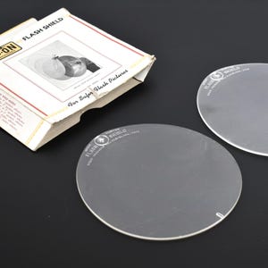 May include: A vintage Clip-On Flash Shield kit with two clear, circular shields and a cardboard box. The box has red and white accents and text that reads "Flash Shield" and "For Safer Flash Pictures."