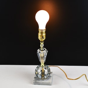 May include: A small, decorative lamp with a clear, faceted crystal base and a gold-colored metal stem. The lamp sits on a square marble base and is topped with a lit light bulb. The cord is a light brown color.
